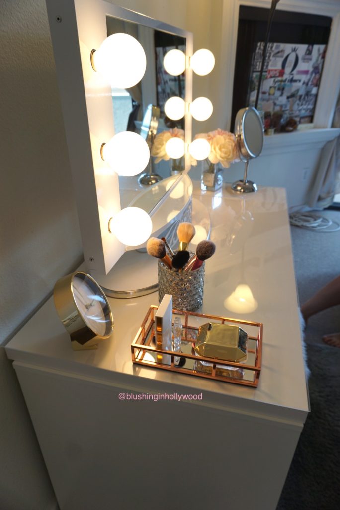 Side view of my vanity where you can see my Vanity Girl Hollywood Lighted Mirror, a rose gold perfume tray, my mercury glass candle holder that I put makeup brushes in, faux peonies, and my Conair magnifying mirror.